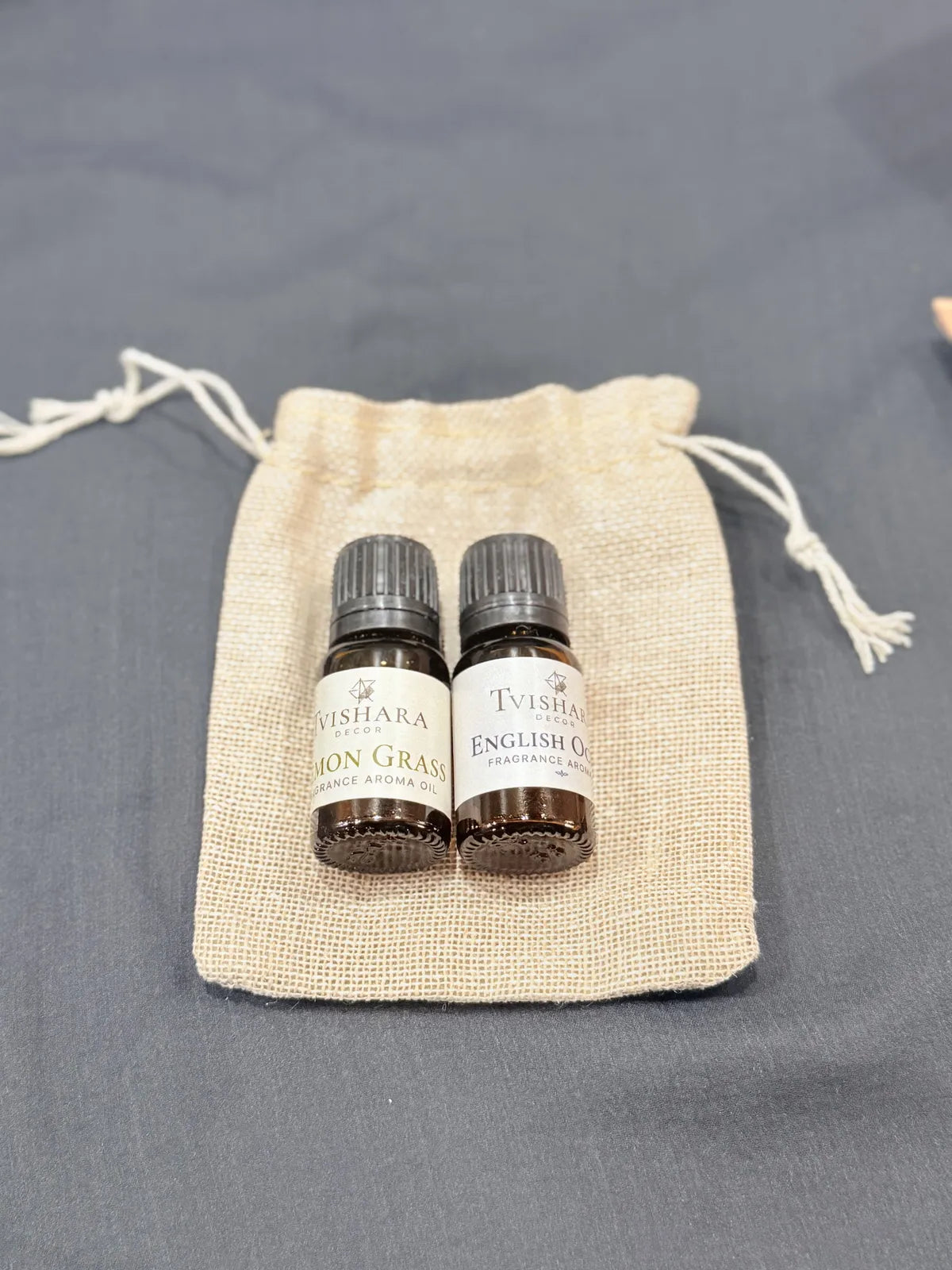 Two small essential oil bottles with white labels and dark caps, placed side by side on a beige drawstring fabric pouch against a gray background.